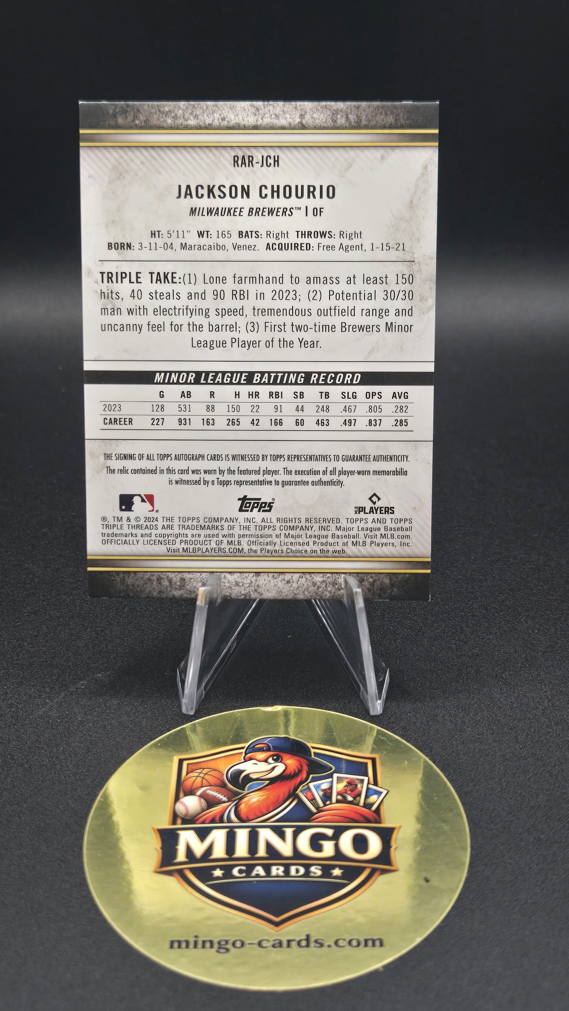 A collectible sports trading card and its circular Mingo Cards display stand on a dark background. The card features baseball player Jackson Chourio with his career stats printed on the back, and the right side shows the round gold-toned Mingo Cards display stand.